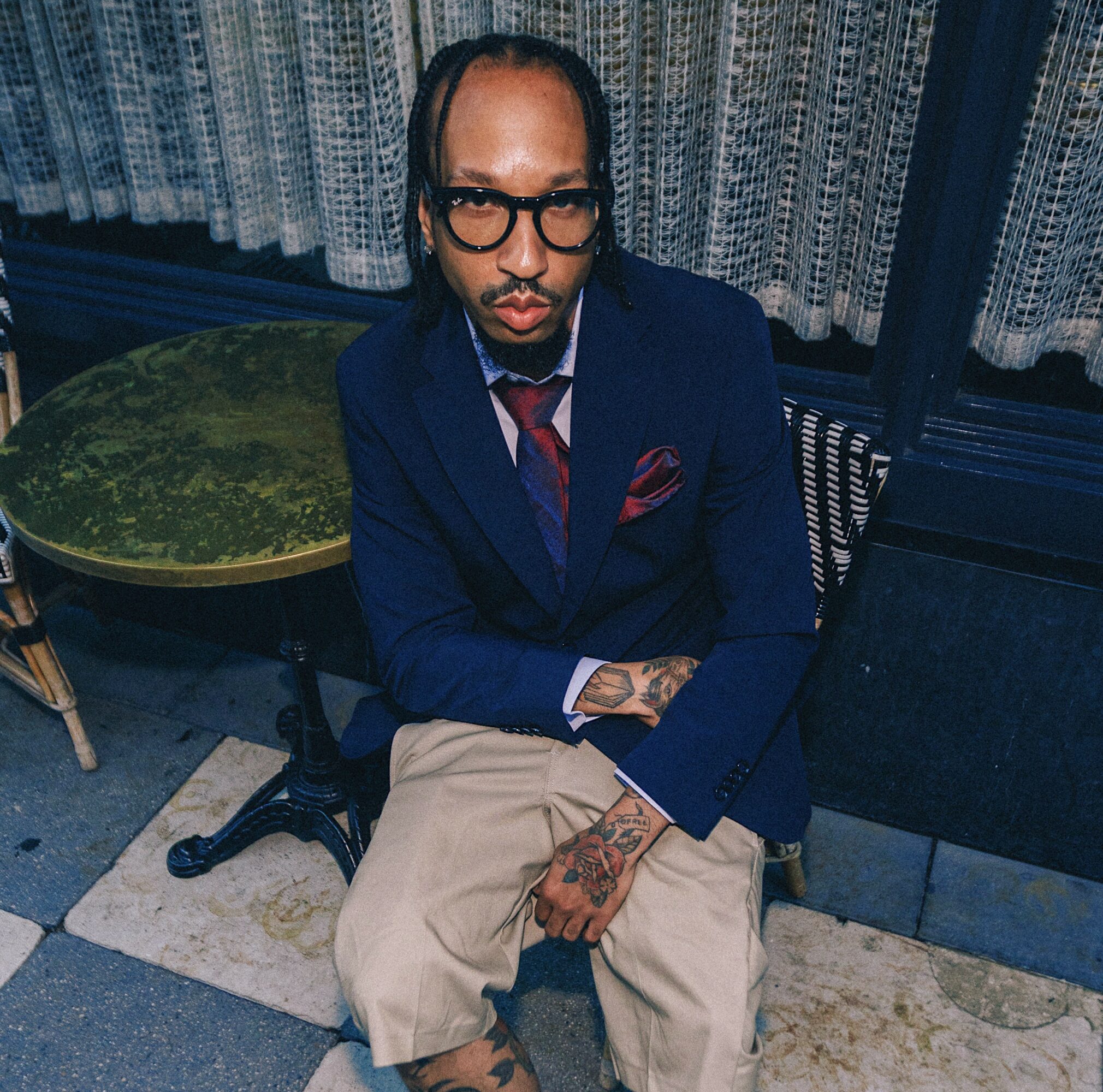 Man with glasses and tattoos sitting outdoors, wearing a navy blazer and beige pants.