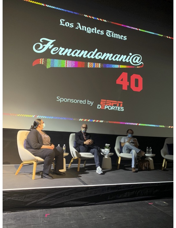 Three people seated on stage in front of a large screen displaying event information, with a dark background and colorful text.