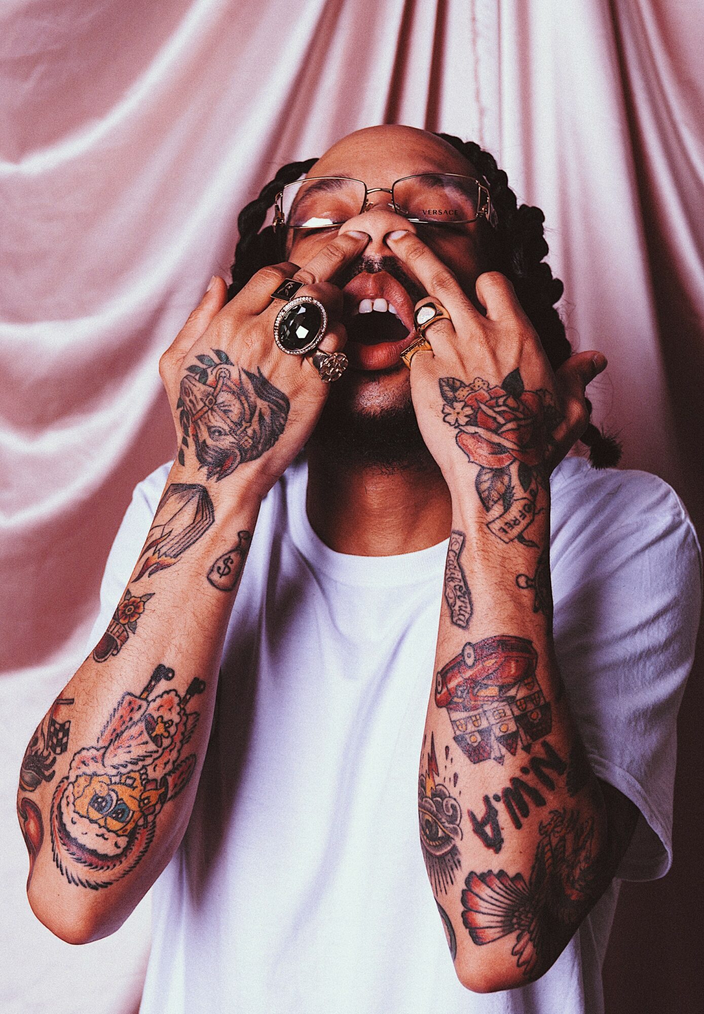 Person with tattoos on arms and hands, wearing glasses, mouth open, hands on face, pink curtain background.