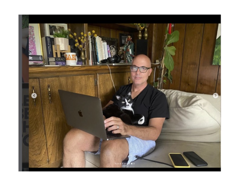 Man sitting on a couch with a laptop, a cat on his lap, bookshelves behind, and two phones on the couch.