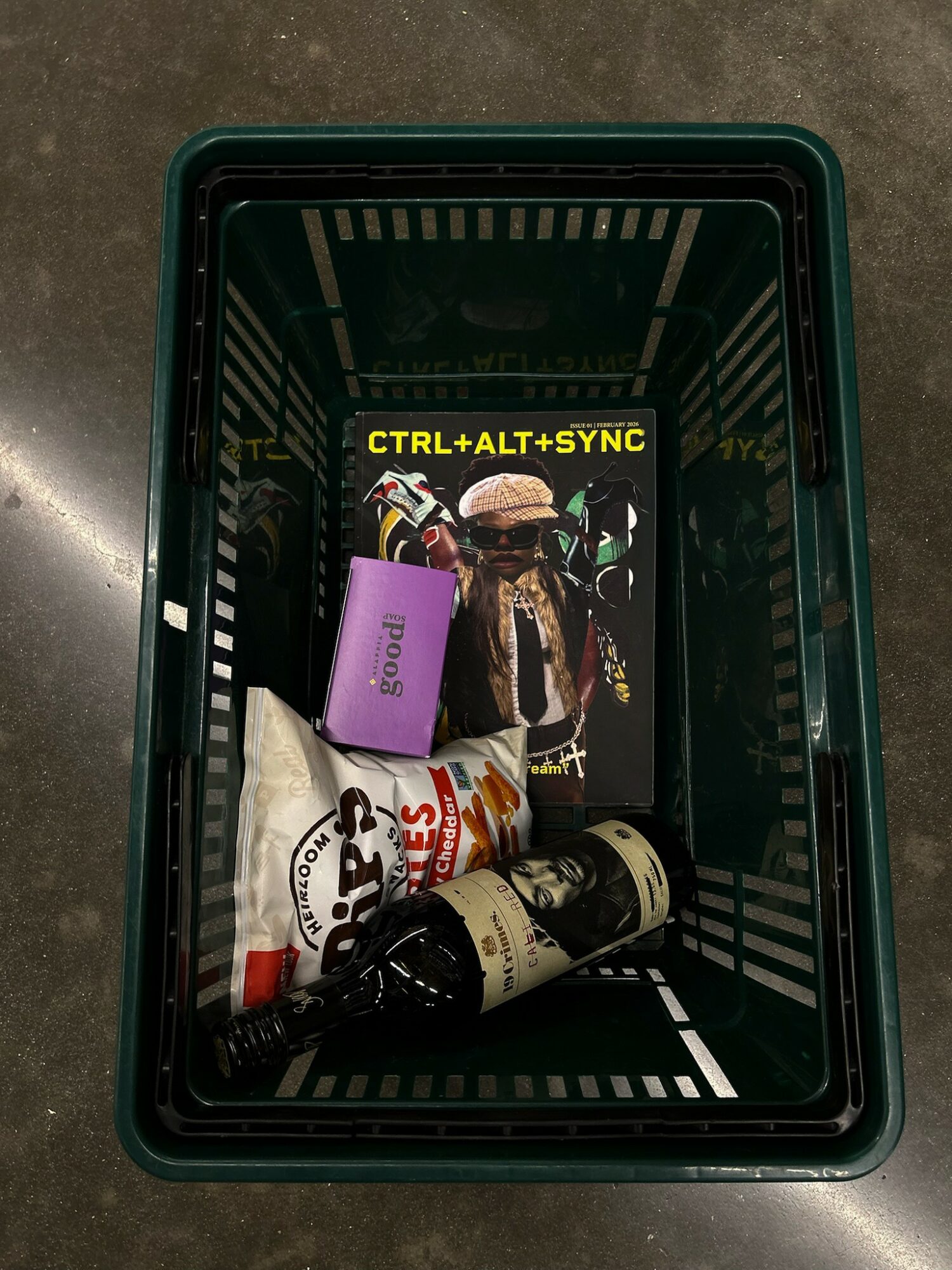 A green plastic basket contains a purple card, a white and red snack bag, a black and white bottle, and a magazine with a person wearing a hat. Text reads 'CTRL+ALT+SYNC'.