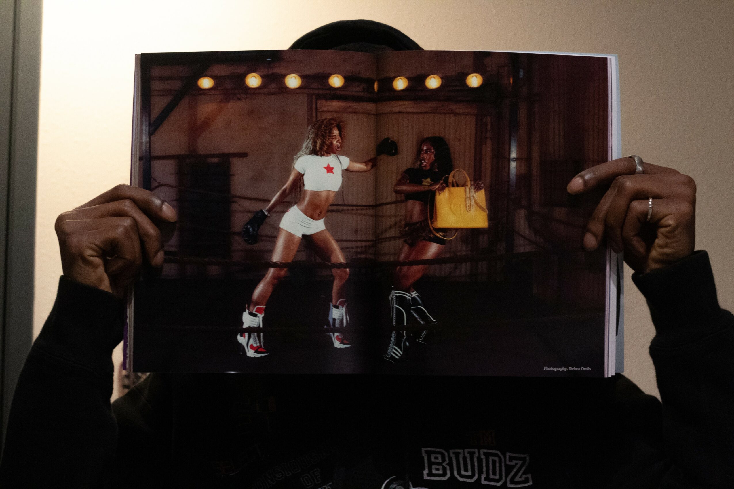 Person holding a photo of two women ice skating indoors, with string lights above. One woman wears a white outfit, the other a yellow bag.