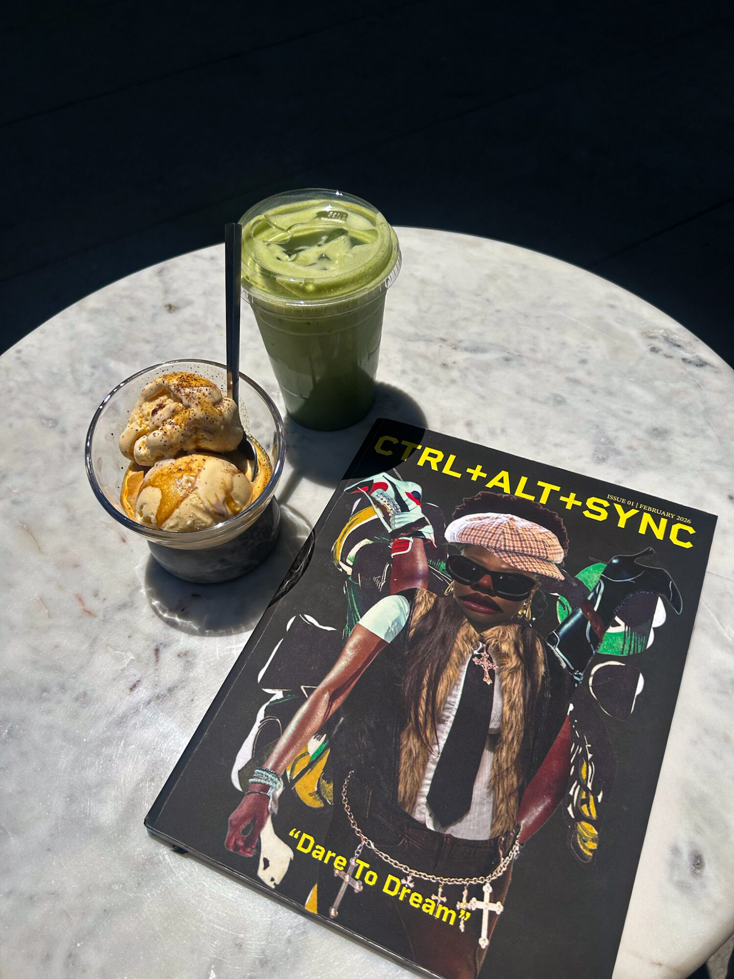 Two beverages on a marble table, one green smoothie and one iced coffee, with a magazine featuring a person in colorful gear.
