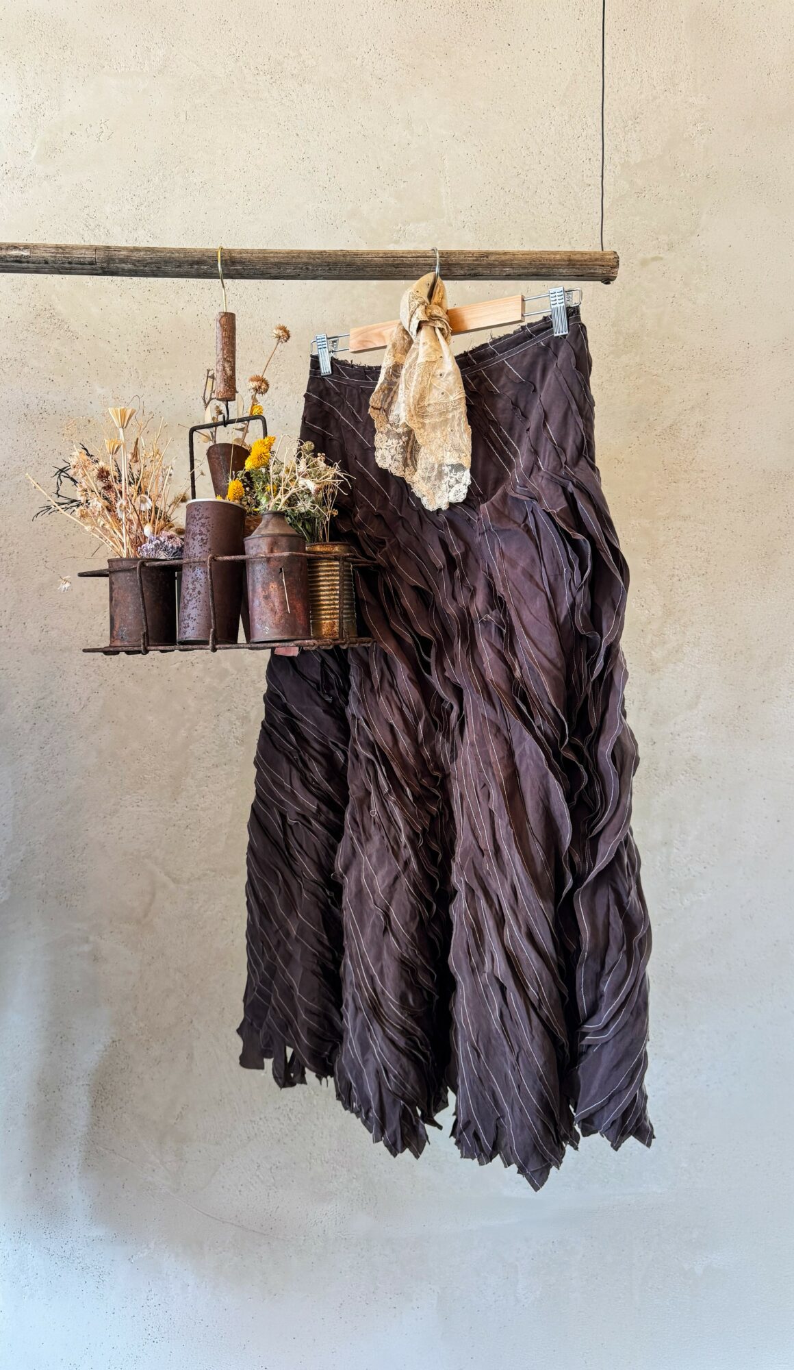 Dark, textured fabric hanging from a wooden rod with dried flowers and a clothespin, against a plain wall.