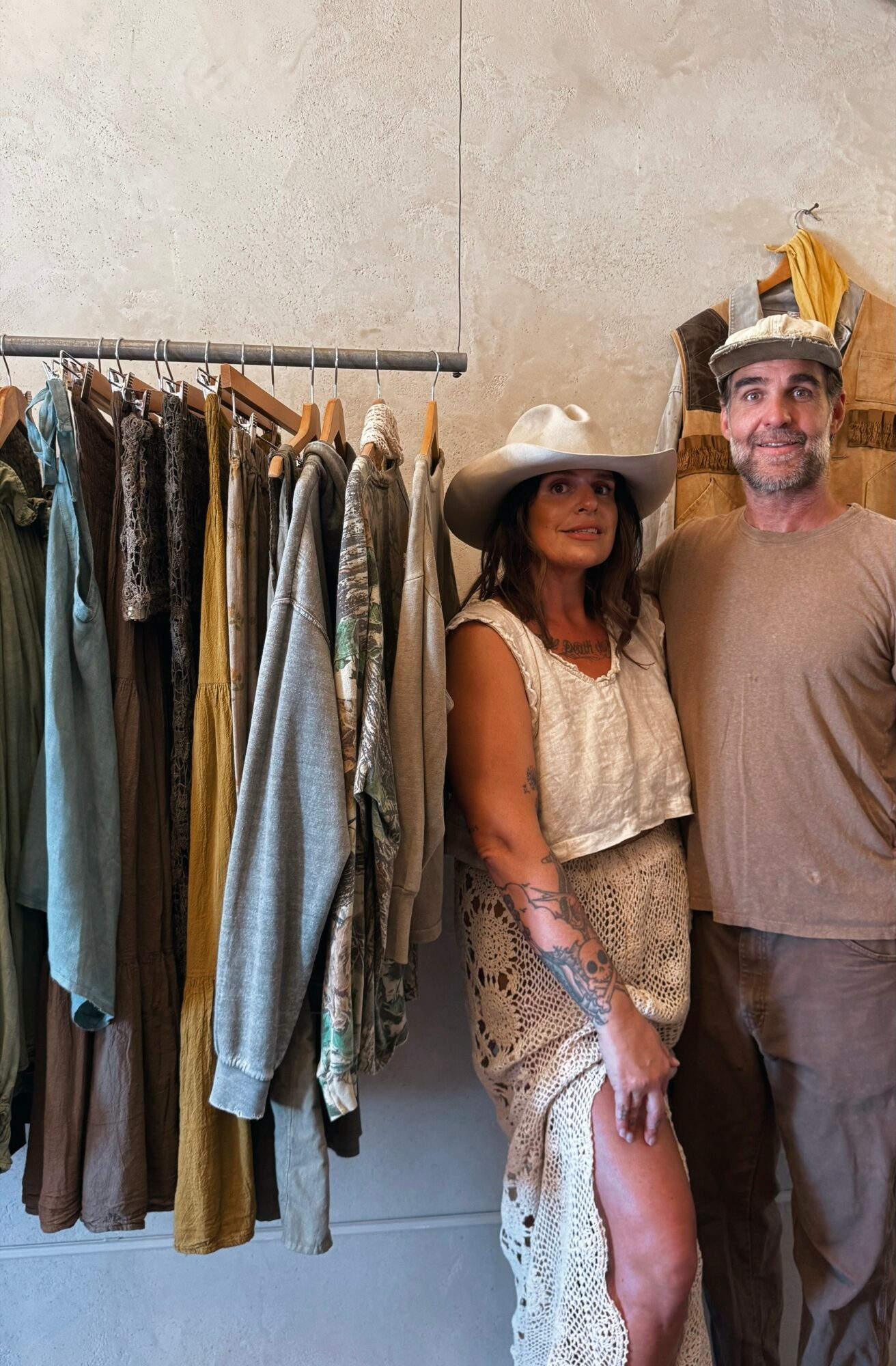 A woman and man stand next to a clothing rack with various garments, in a neutral-colored room.