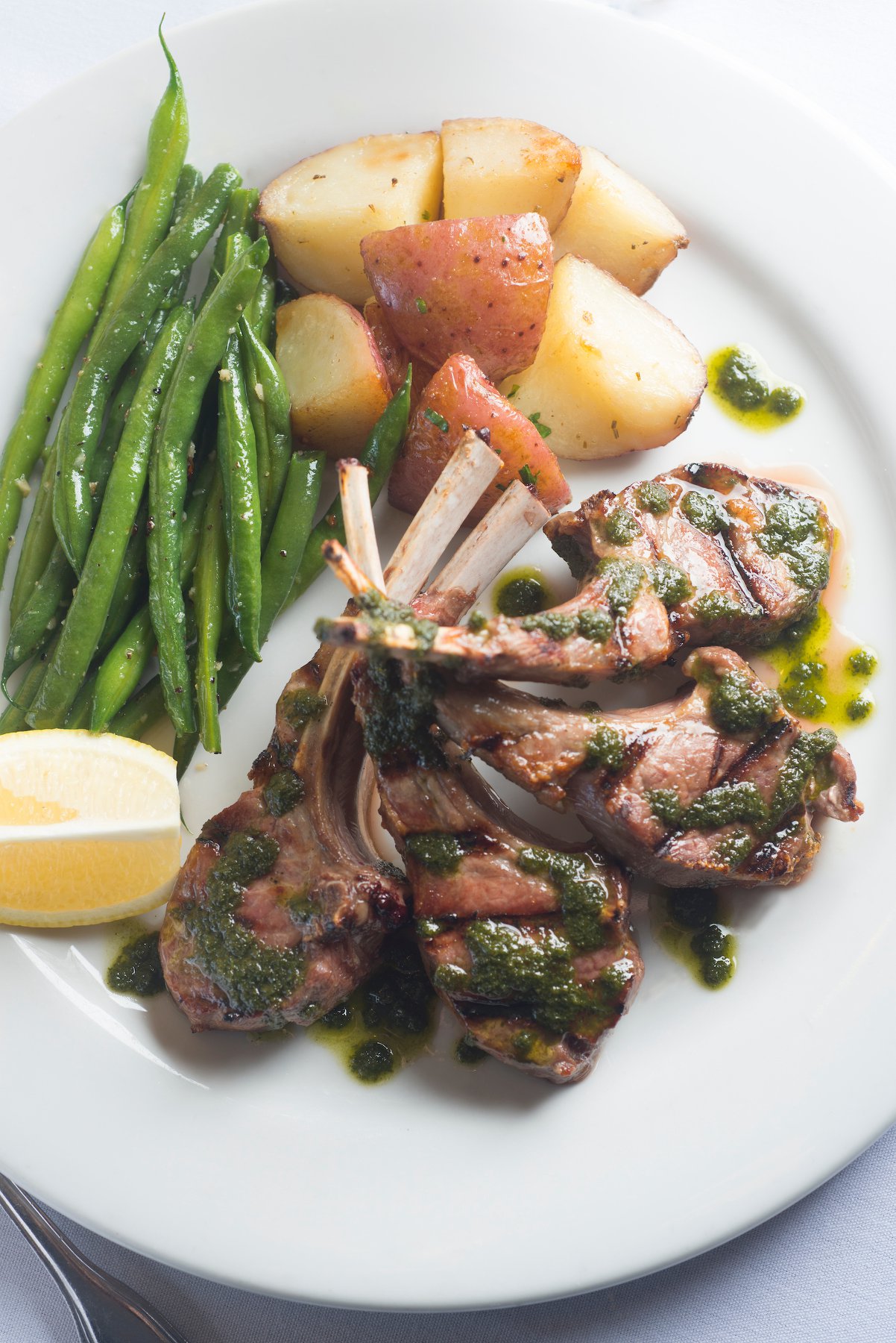 Plate with green beans, roasted potatoes, and grilled lamb chops drizzled with green sauce, lemon wedge on side.