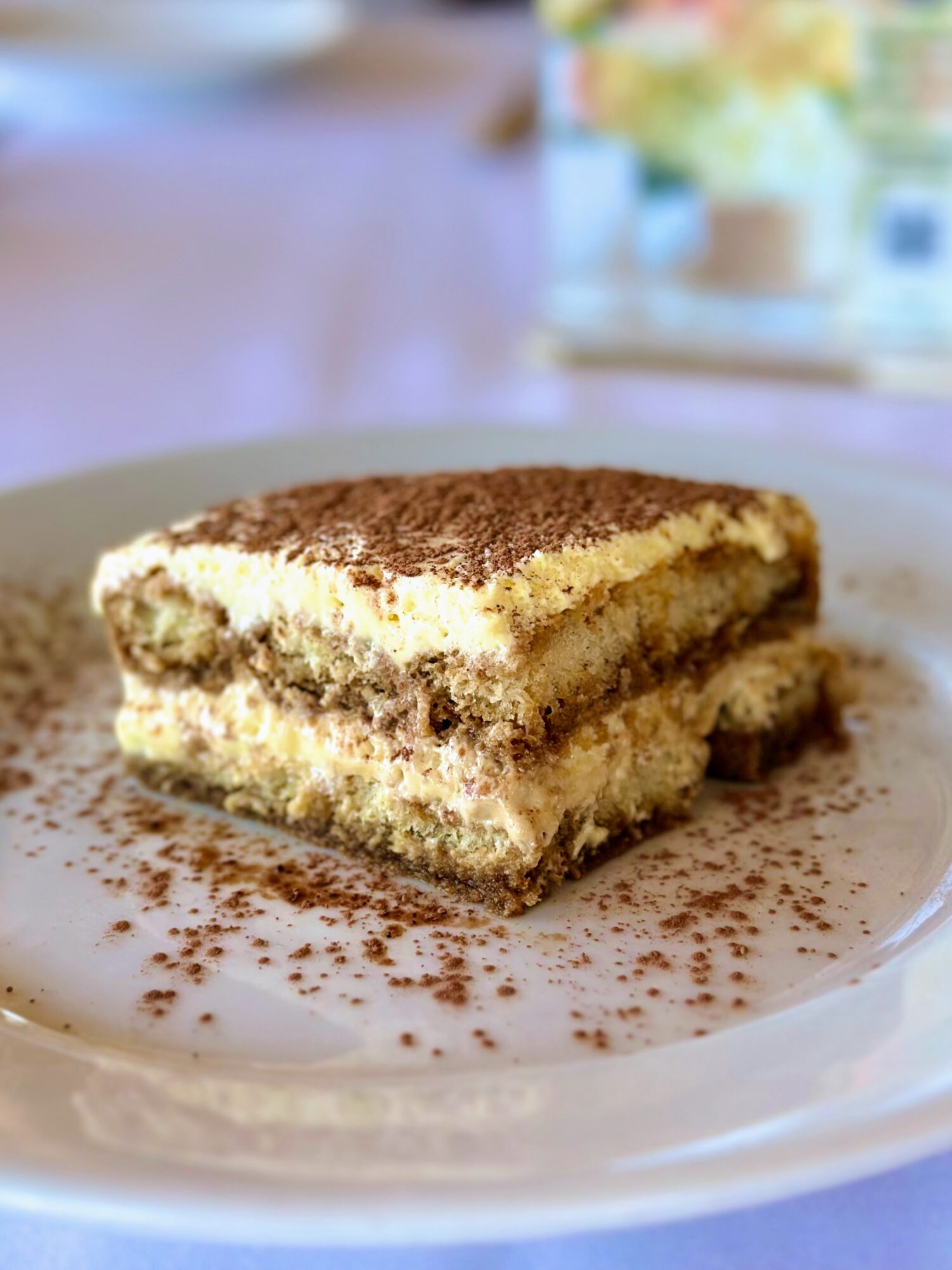 A slice of layered tiramisu dessert with cocoa powder on top on a white plate, garnished with cocoa powder around it.
