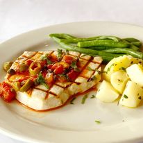 Grilled fish fillet topped with tomato-based sauce, served with green beans and potato salad on a white plate.