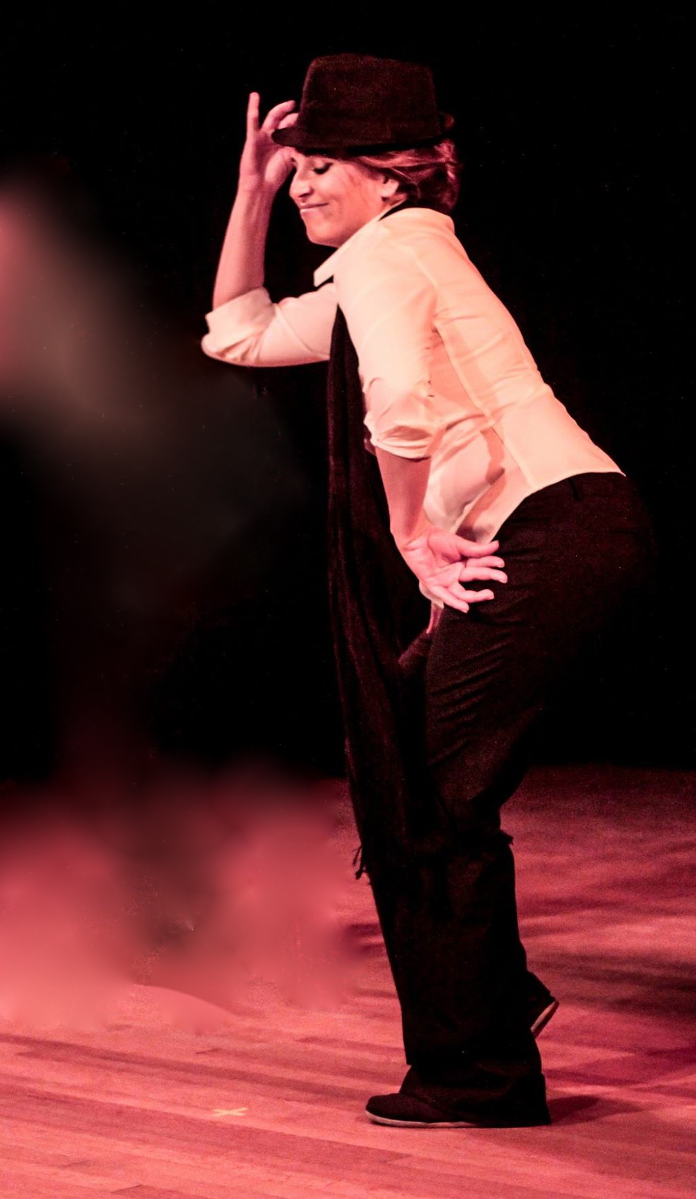Person with short hair, wearing a hat, beige top, and black pants, posing with one hand on hip and head tilted.