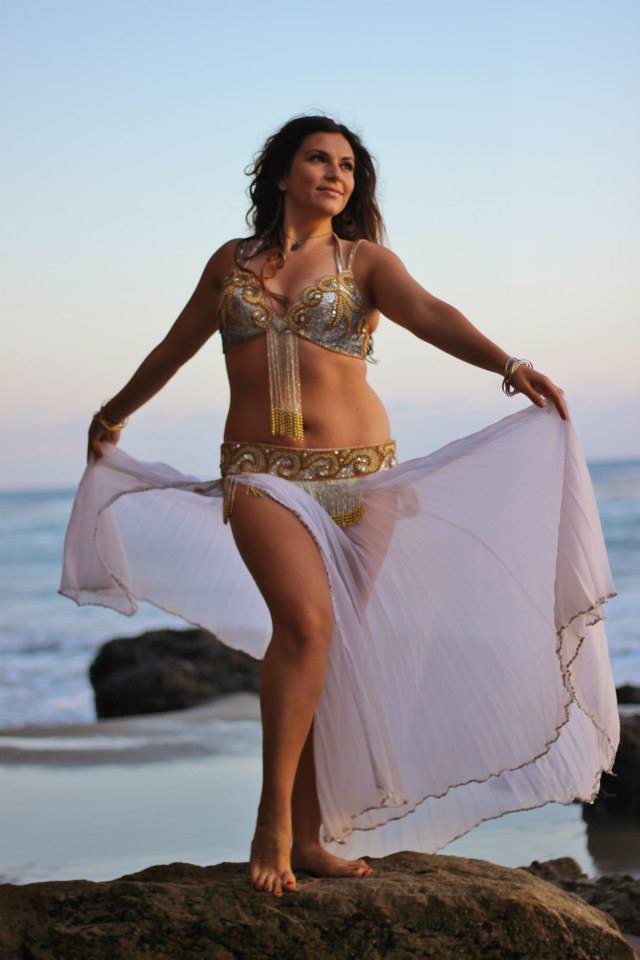Woman in a belly dance costume standing on a rock at the beach, holding a sheer white fabric, with ocean and sky in background.