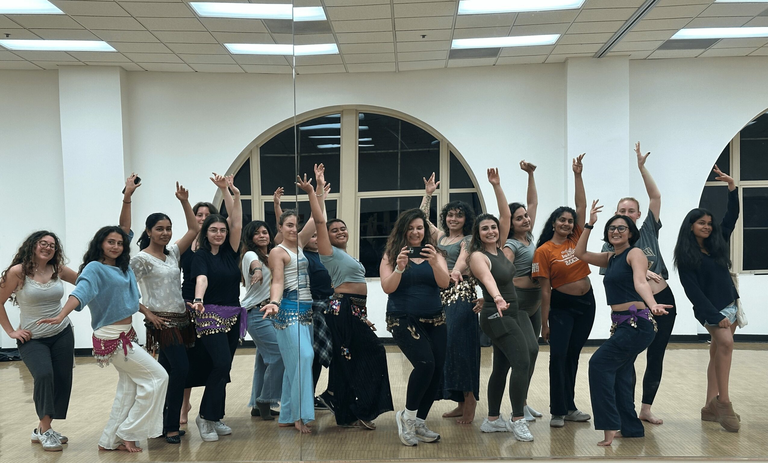 Group of women dancing indoors, raising arms, smiling, in a spacious room with large arched windows.