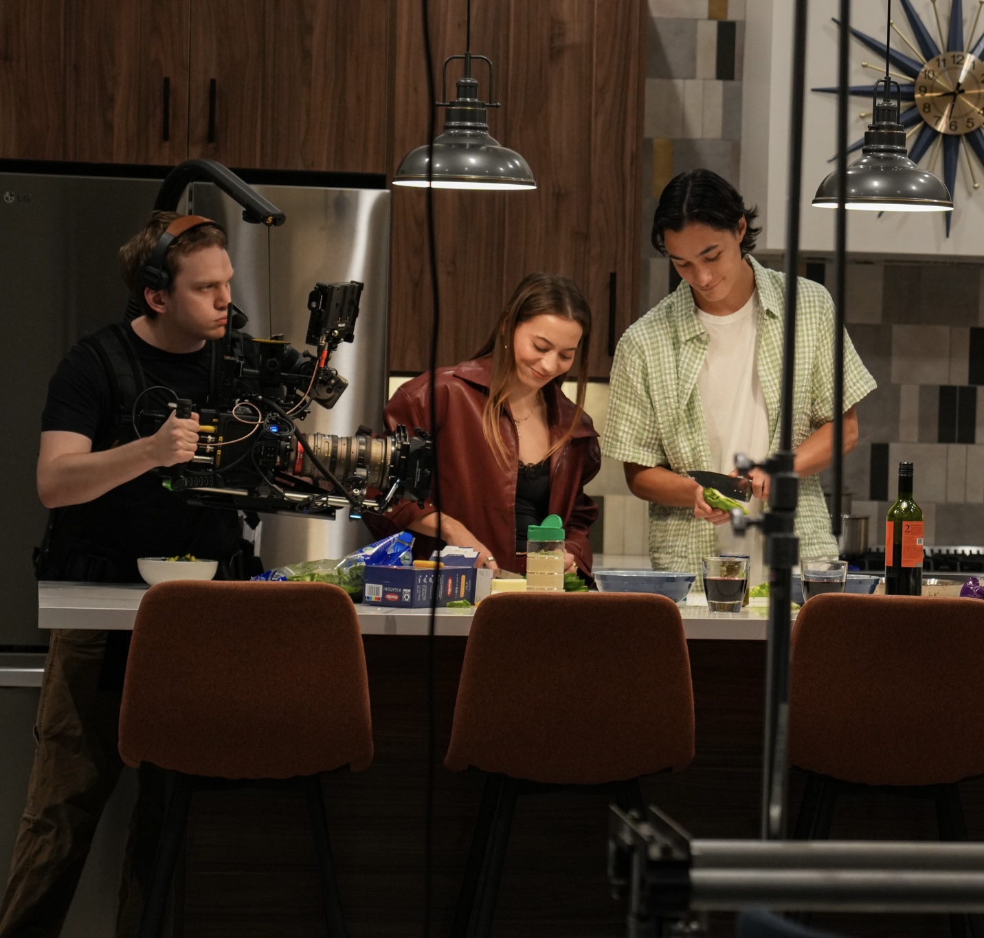 Three people in a kitchen with a camera operator filming, two women and one man preparing food at a counter.