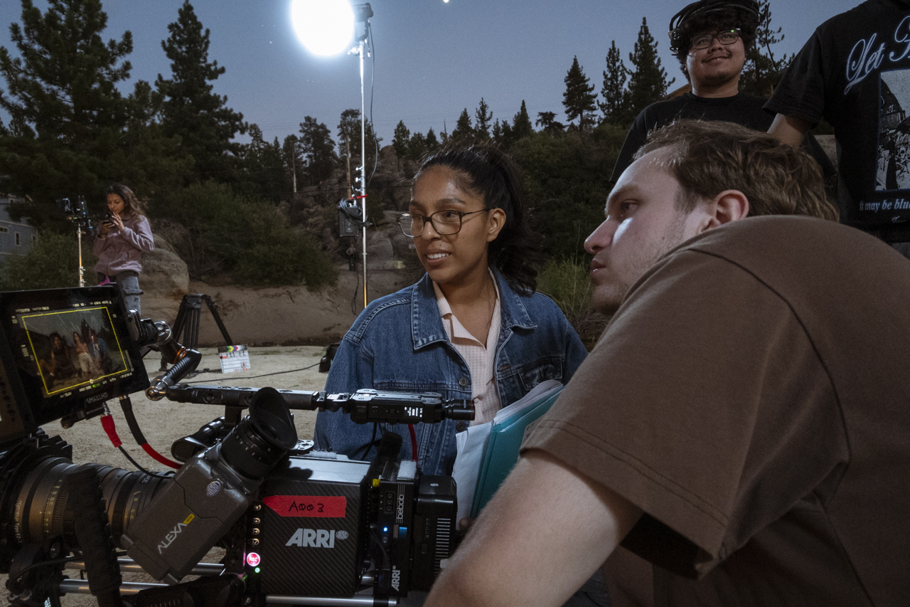 Group of people outdoors at night, with a camera and equipment, some smiling and talking, trees in the background.