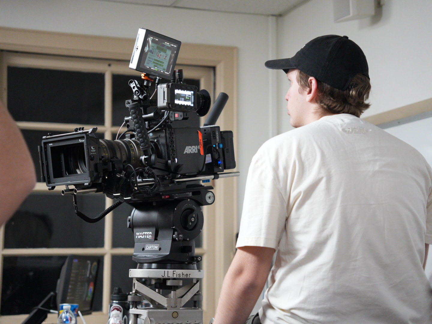Person in white shirt and black cap operating a professional video camera indoors, with a window in the background.