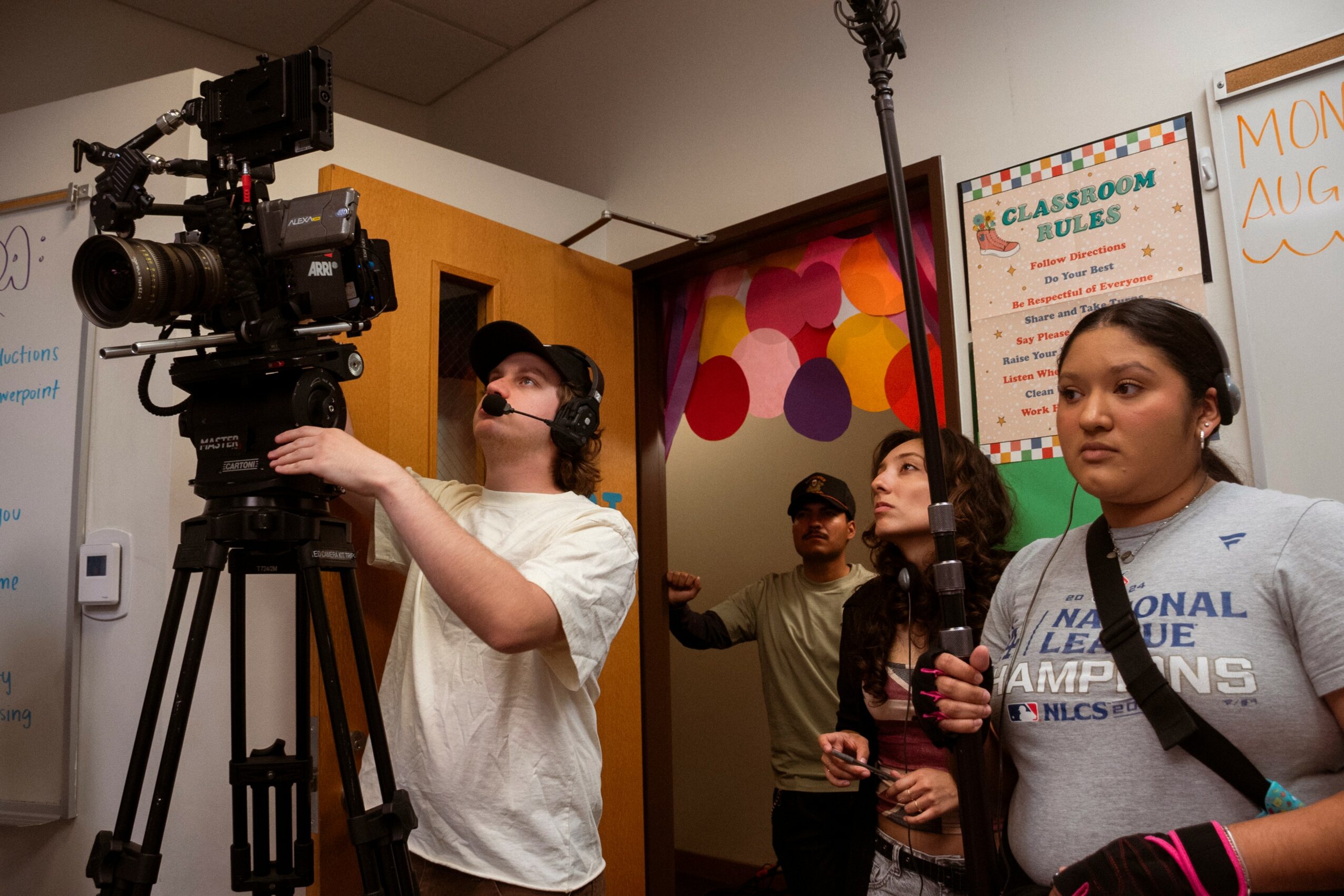 Group of children and a camera operator in a classroom, with colorful wall art and a whiteboard in the background.