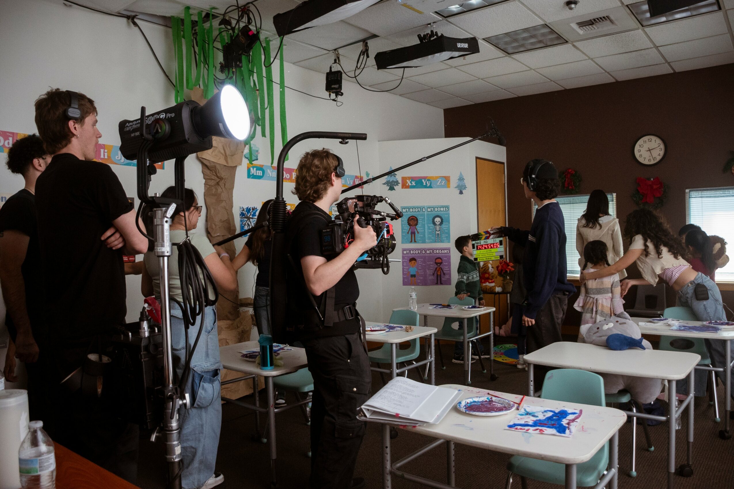 Group of people in a classroom with cameras and lighting equipment, some students at desks, a teacher at the front.