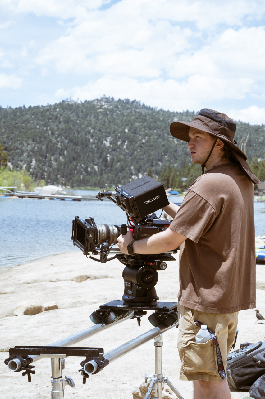 Person operating a professional camera on a tripod near a body of water with a mountain in the background, wearing a hat and casual clothing.