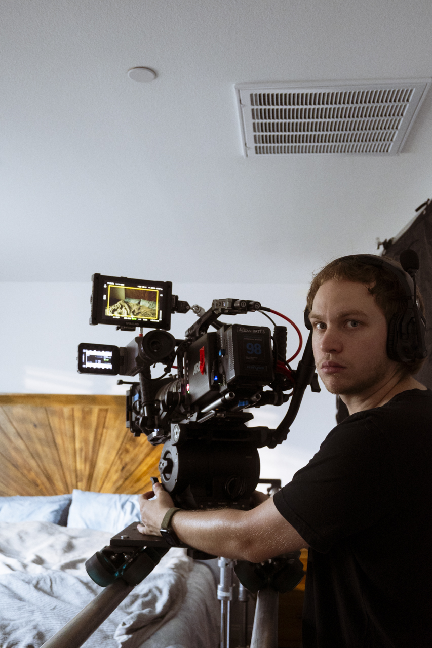 Person operating a professional camera on a tripod in a room with a bed and wooden headboard, wearing headphones.