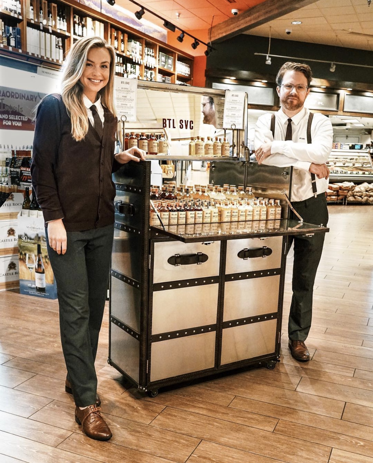 Two people stand behind a display cart in a store, smiling, with shelves of products in the background.