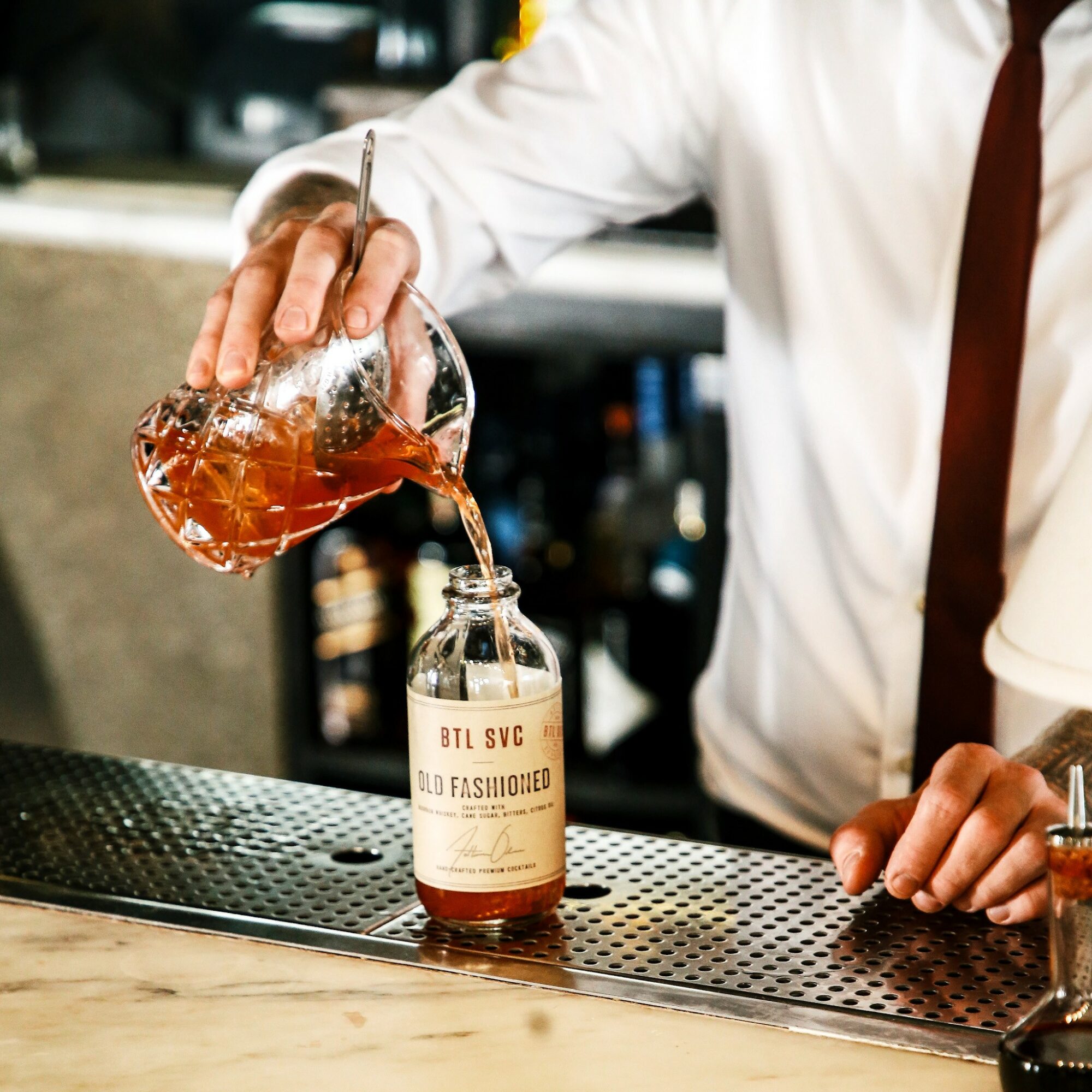 Person pouring a drink from a glass decanter into a bottle on a bar counter, wearing a white shirt and brown tie.