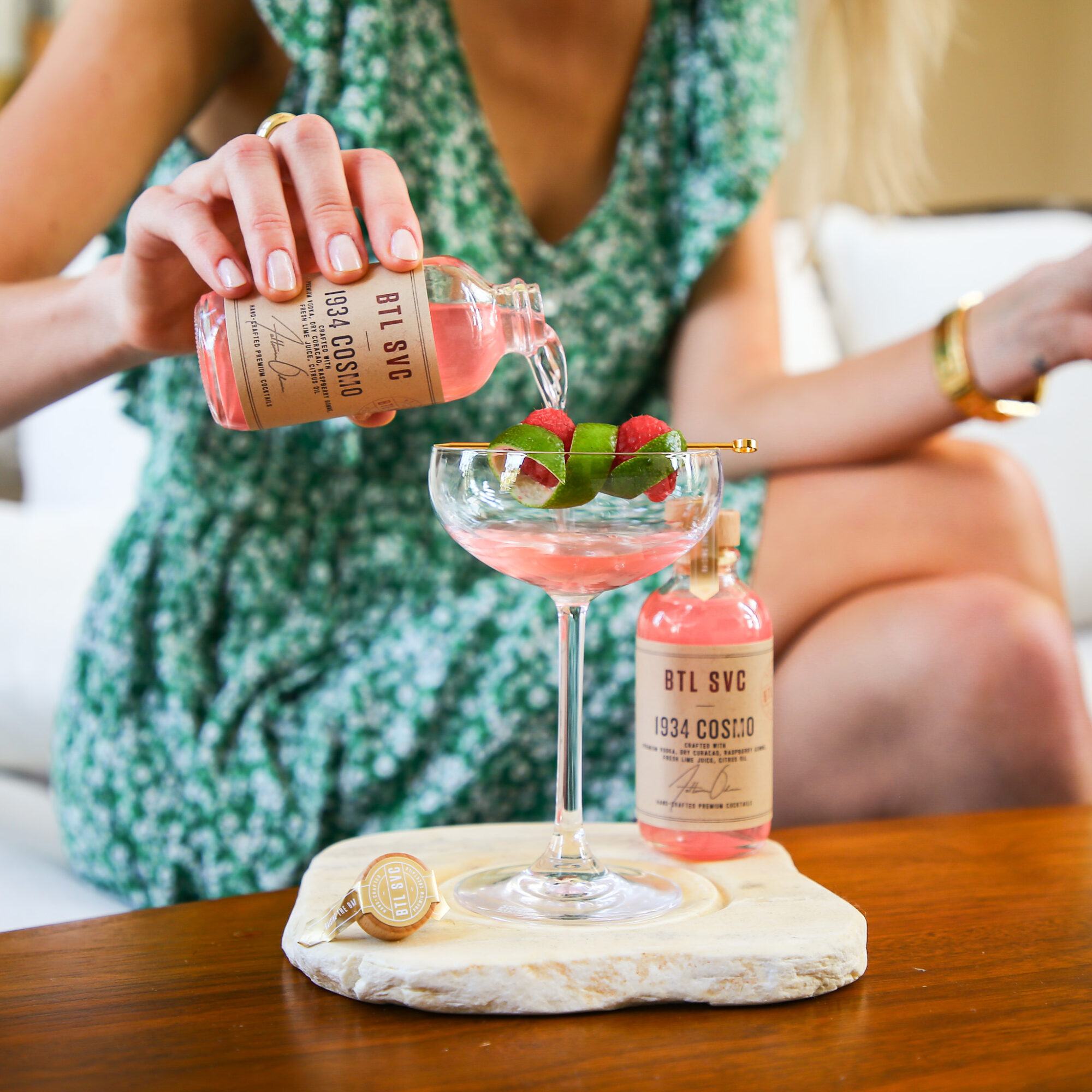 Person pouring pink drink into a glass with strawberries, on a marble slab, with a small bottle nearby, in a casual setting.