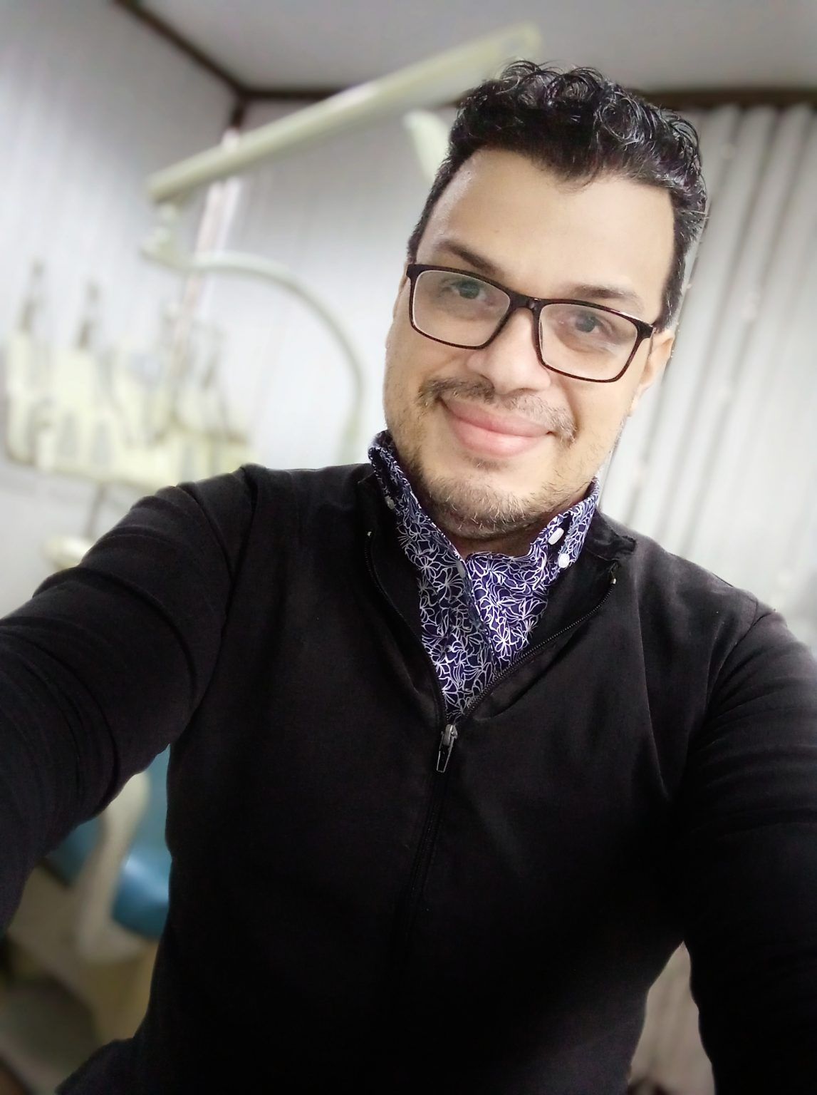 Young man with glasses smiling, wearing a black jacket and patterned shirt, in a room with medical equipment.