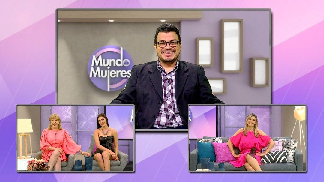 Three people on a TV set with a Mundo Mujeres logo, two women sitting on couches, one man standing behind a desk.