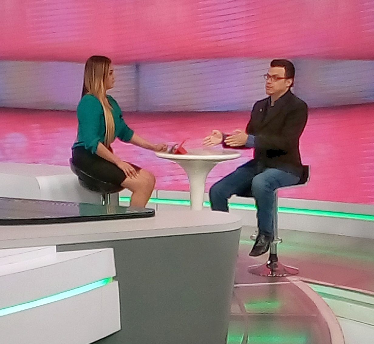 Two people sit at a round table on a TV set with pink and green background, engaging in conversation.