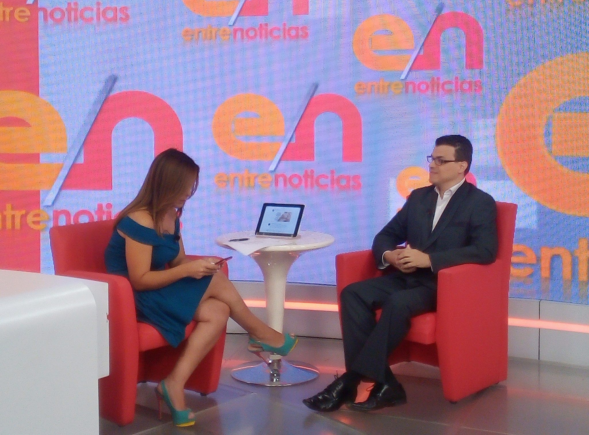 A woman and a man sit in red chairs on a TV set with a colorful background, a small table with a laptop between them.