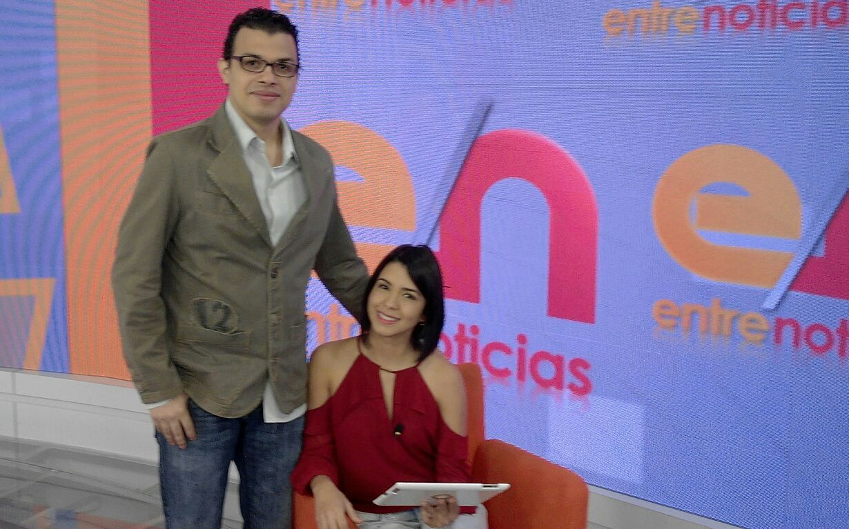 Man and woman in a studio with a colorful background, woman seated with a tablet, man standing beside her.