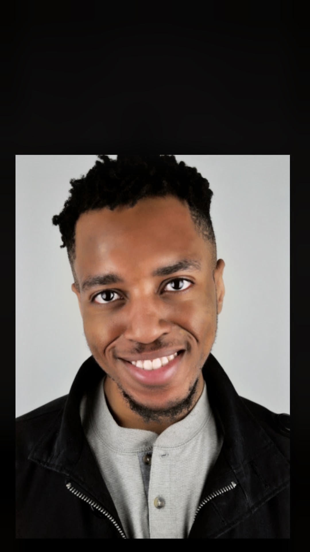 Smiling young man with short curly hair, wearing a light gray shirt and black jacket, against a plain background.