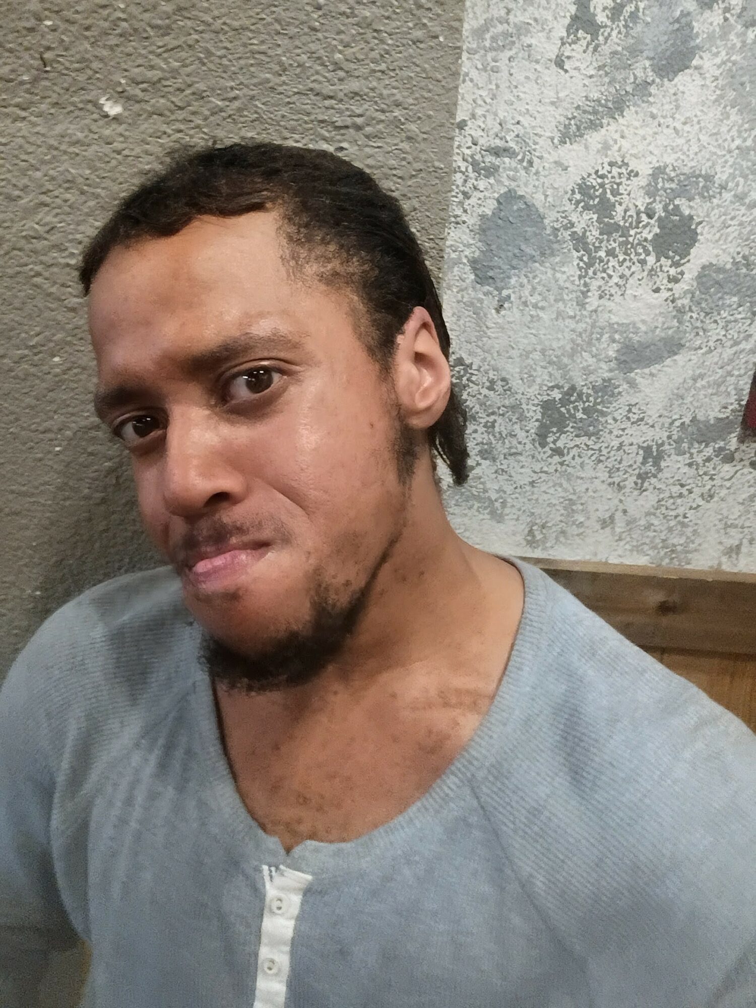 Young man with short curly hair and a beard, wearing a light gray shirt, sitting against a textured wall.