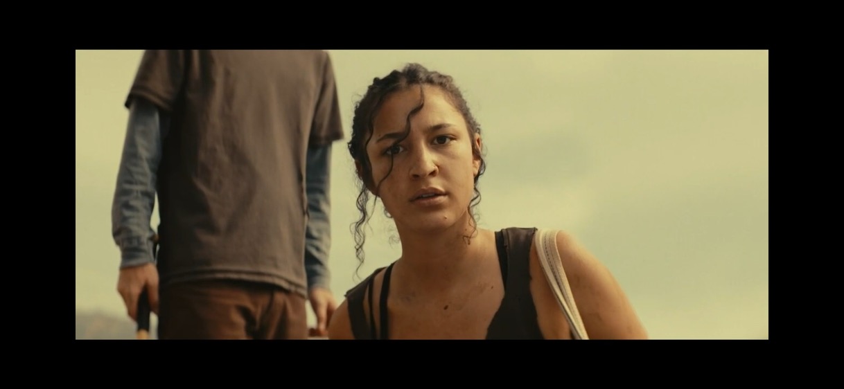 A woman with curly hair looks concerned, with a person standing behind her, against a cloudy sky.