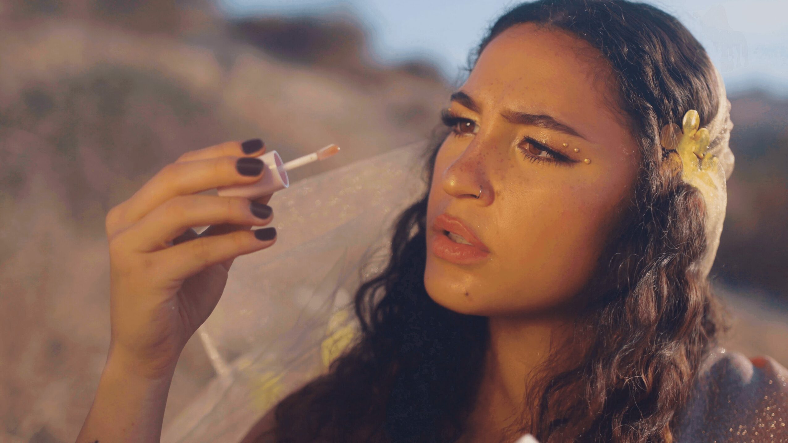 Woman with long dark hair holding a cigarette outdoors, looking thoughtful, with a blurred natural background.