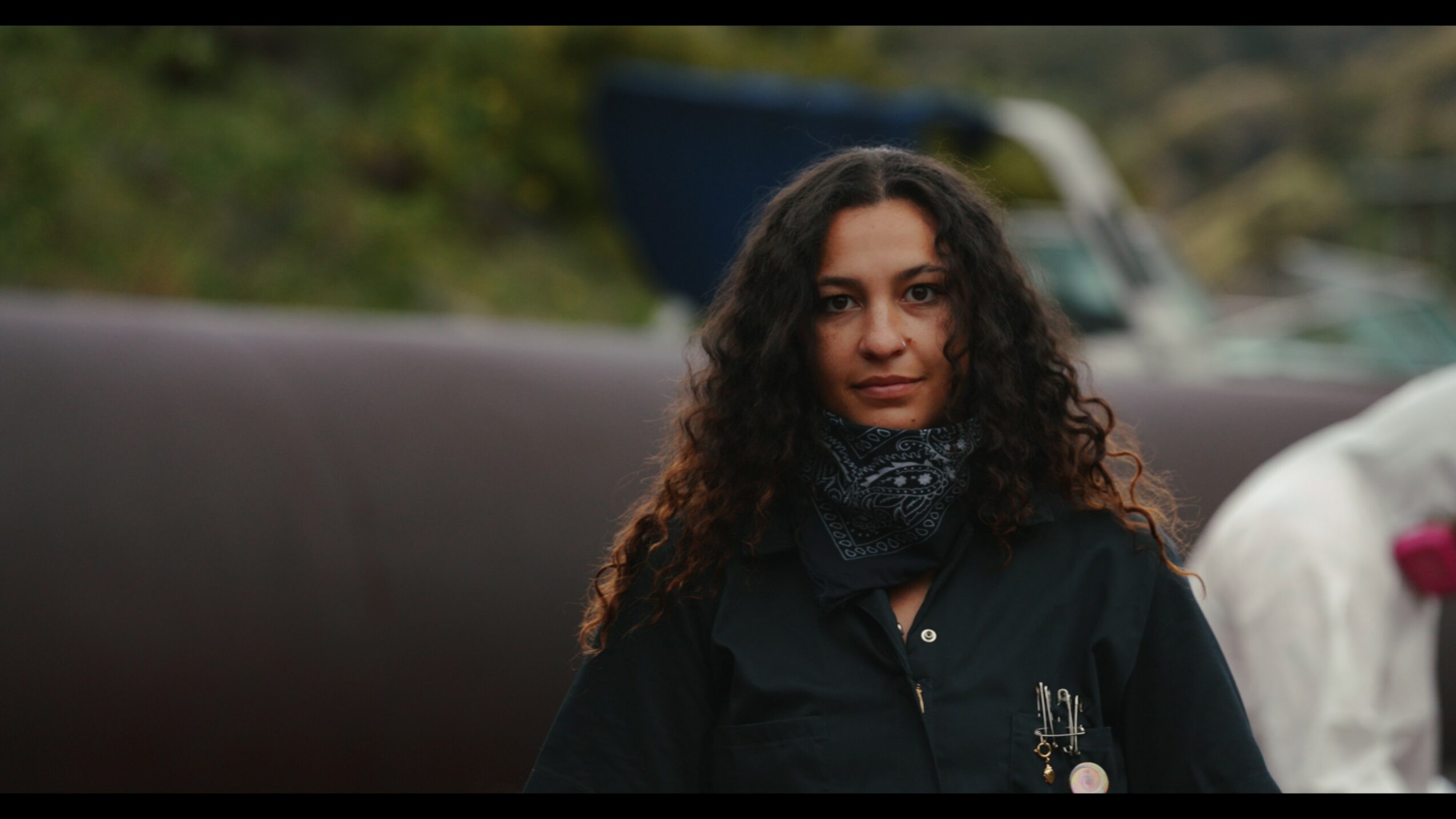 Woman with curly hair wearing a dark jacket and scarf outdoors, blurred background with greenery and vehicles.