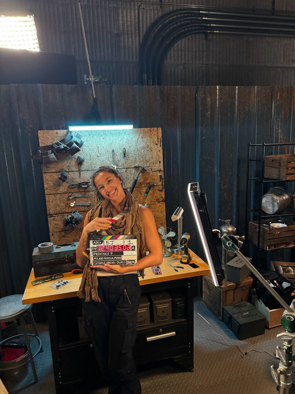 Woman standing behind a desk in a studio, holding a colorful book, with equipment and a mirror in the background.