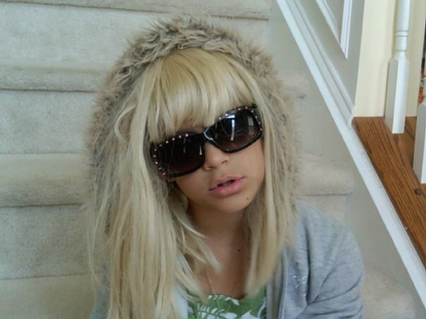 Child with long blonde hair wearing large sunglasses and a furry hat, sitting on stairs indoors.