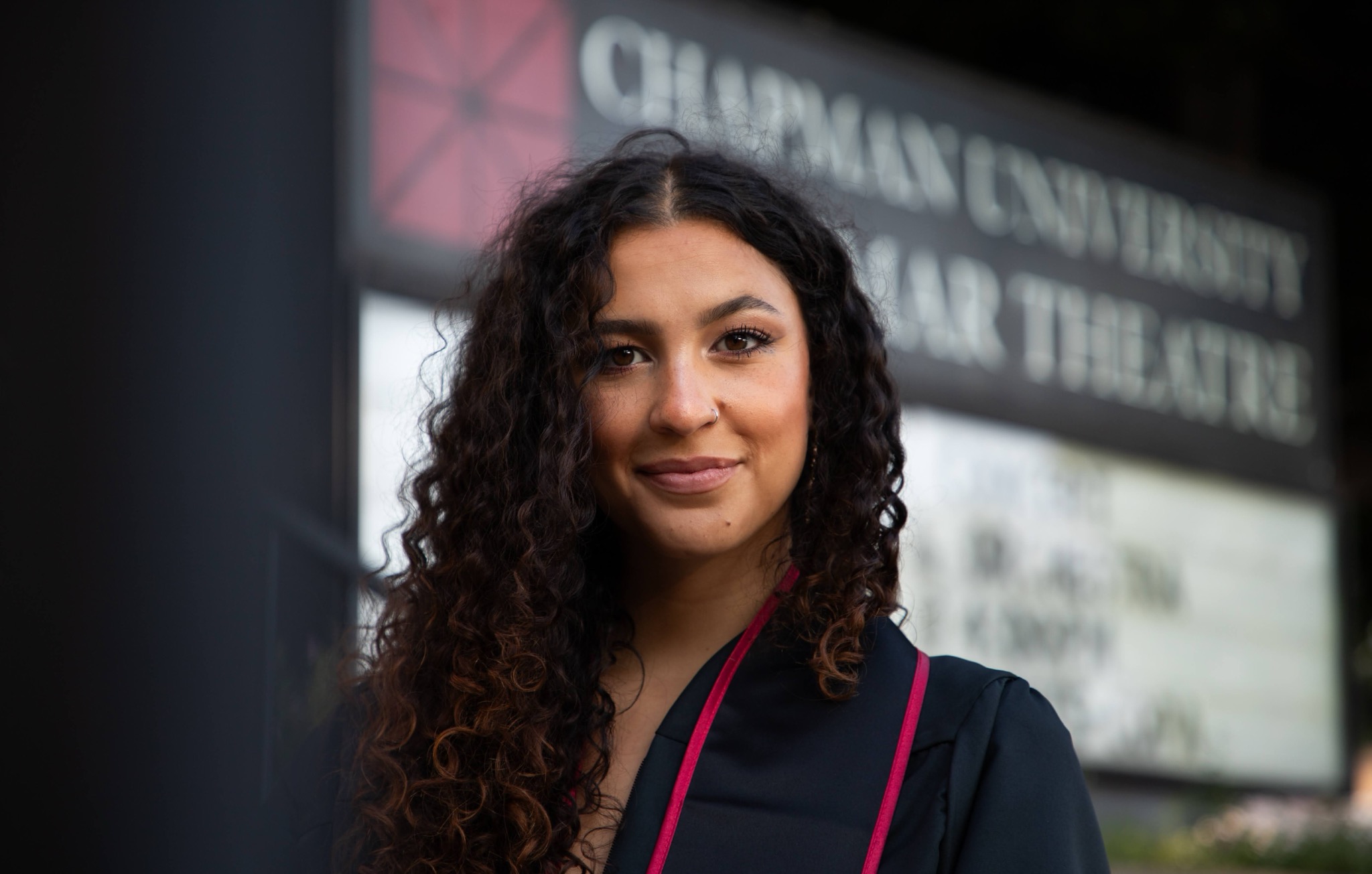 Young woman with curly hair smiling outdoors in front of a sign for a university theater.