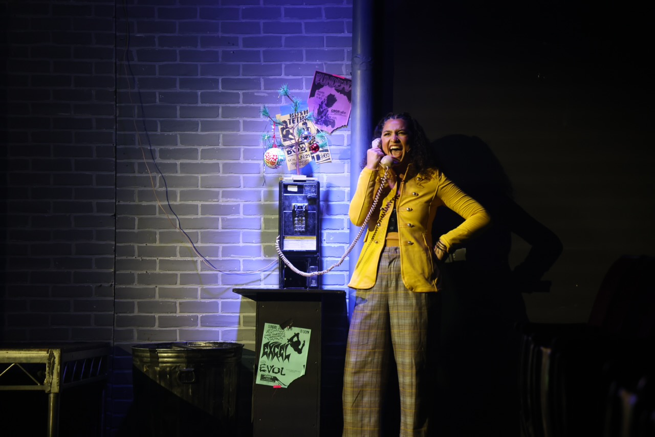 Woman in yellow jacket smiling and speaking into a microphone, standing next to a vending machine against a brick wall.