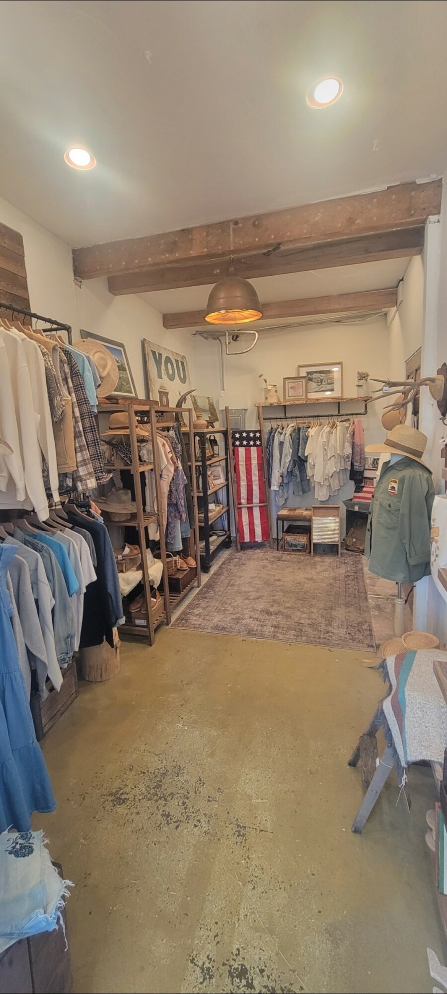 Clothing store interior with racks of clothes, shelves, and a wooden beam ceiling, viewed from the entrance.