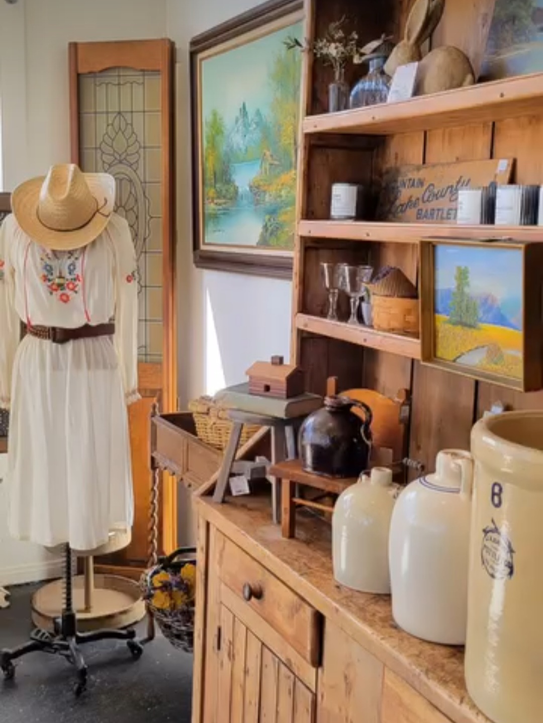 Display of vintage items including a dress on a mannequin, paintings, and ceramic jars on wooden furniture.