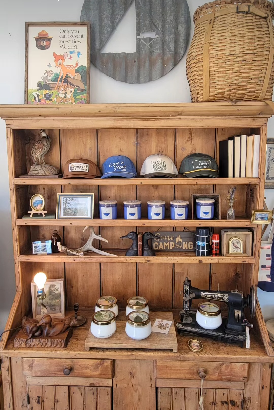 Wooden shelf with books, hats, jars, and camping items, topped with a mirror, basket, and framed picture, against a gray curtain.