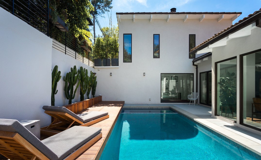 Modern house with a swimming pool, outdoor lounge chairs, and tall cacti on the left, under a clear sky.