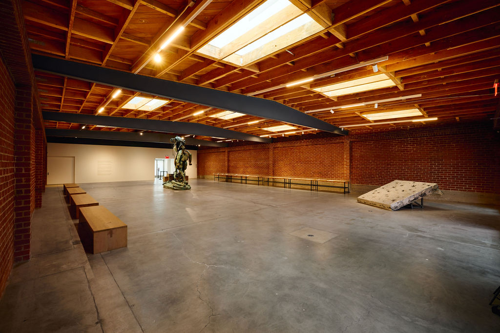 Empty indoor space with brick walls, wooden ceiling, benches, and a sculpture in the background.