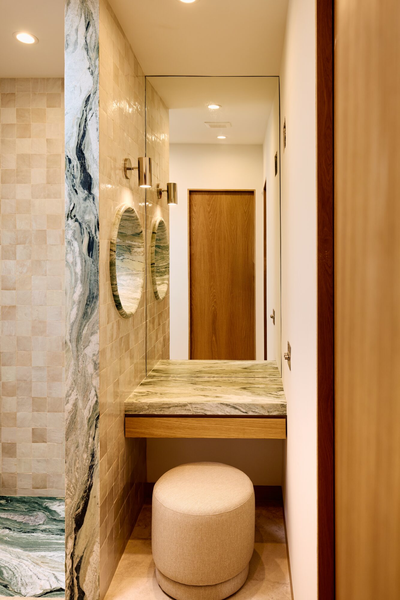 Narrow bathroom with a marble countertop, round mirror, and a beige ottoman, with a wooden door at the end. Word count: 20