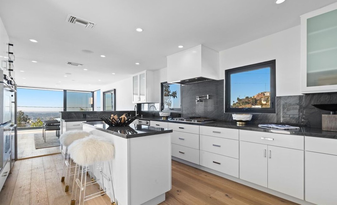 Modern kitchen with white cabinets, black countertops, large windows, and a view outside, with a kitchen island and bar stools.