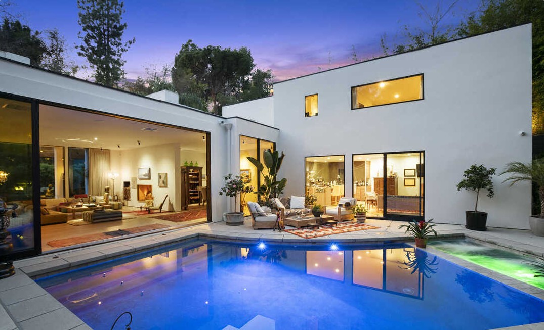 Modern house with large glass windows and a swimming pool in the backyard, trees in the background, evening sky, and outdoor seating area.