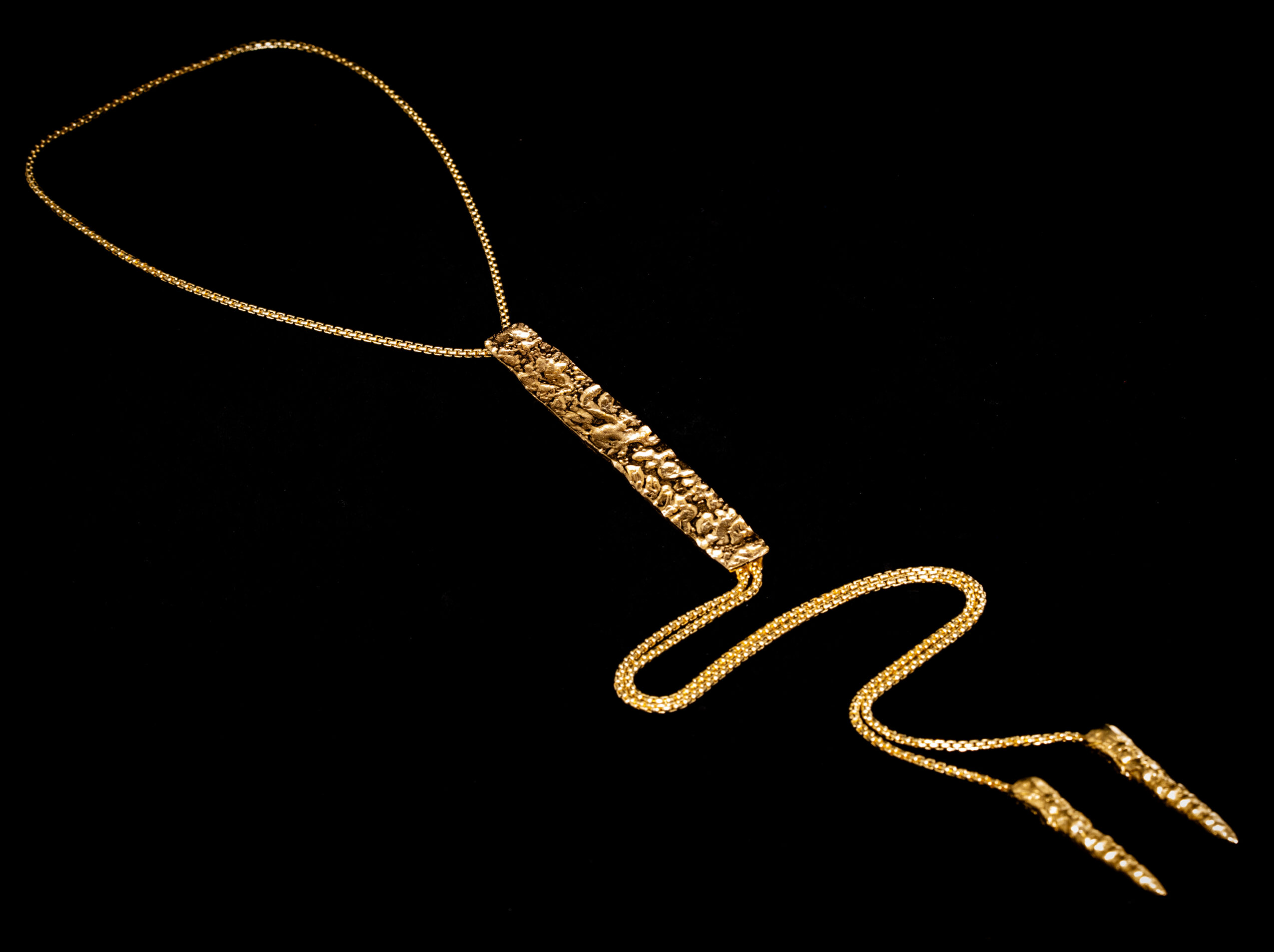 Gold necklace with a pendant shaped like a question mark and a pointed end, against a black background.