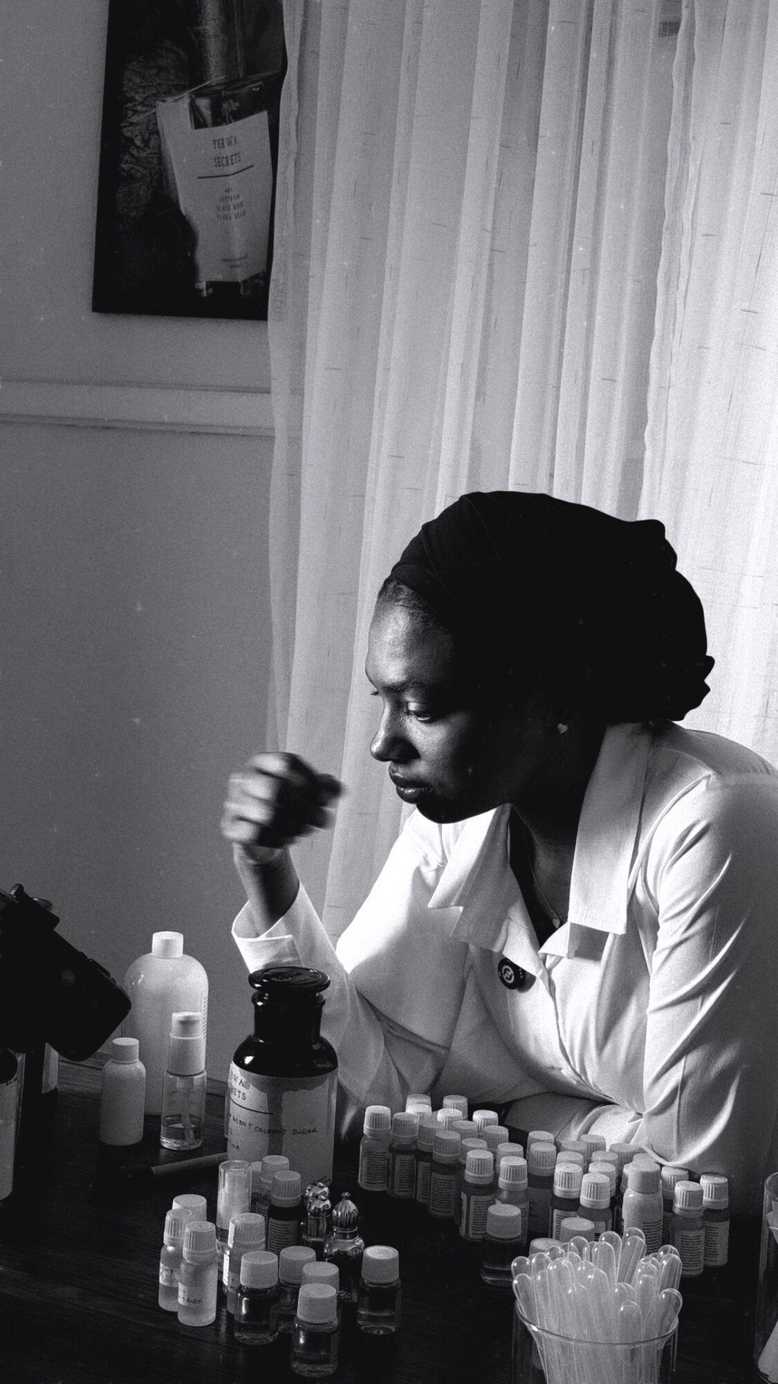 Woman with braided hair in a white coat sitting at a table with bottles and containers, looking down thoughtfully.