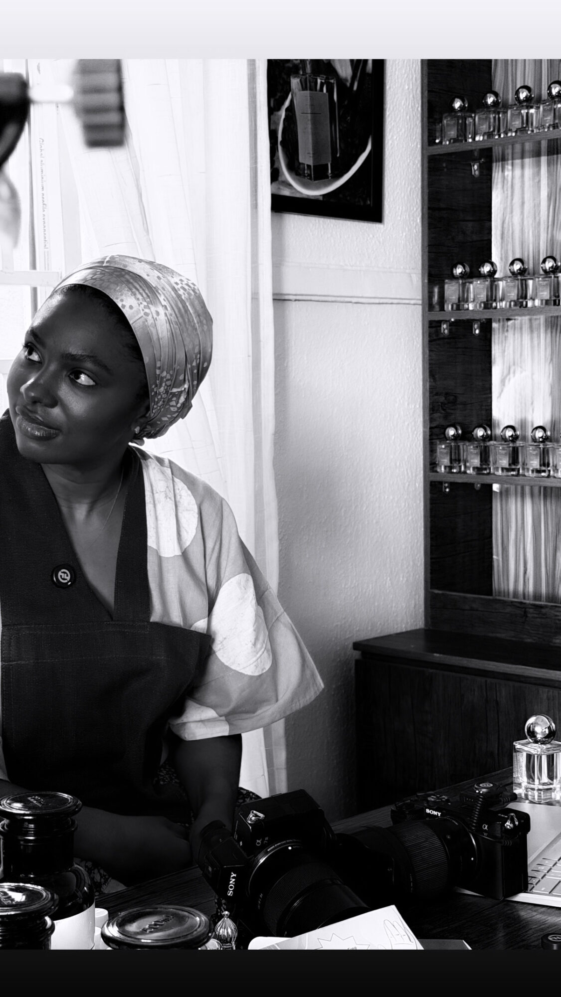 Woman wearing a headscarf sitting in a room with cameras and a display cabinet of bottles.